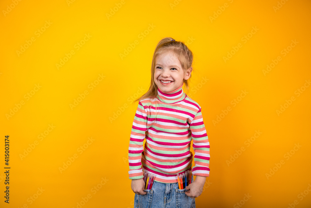 Laughing child in a striped sweater standing on a yellow background with felt-tip pens in jeans pockets Laughing child in a striped sweater standing on a yellow background with felt-tip pens in jeans pockets