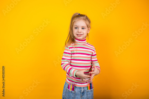Young girl artist in a striped sweater standing on a yellow background with felt-tip pens in jeans pockets