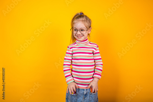Funny little girl with glasses in a striped jacket standing on a yellow background with markers in the pockets of her jeans