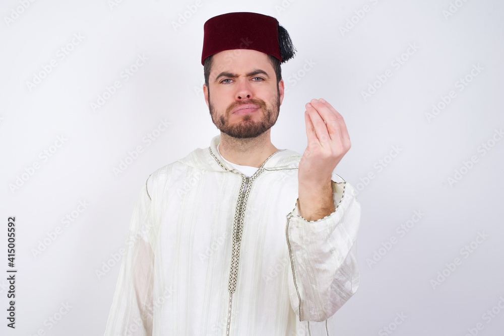 young handsome Caucasian man wearing Arab djellaba and Fez hat over white wall angry gesturing typical italian gesture with hand, looking to camera young handsome Caucasian man wearing Arab djellaba and Fez hat over white wall angry gesturing typical italian gesture with hand, looking to camera