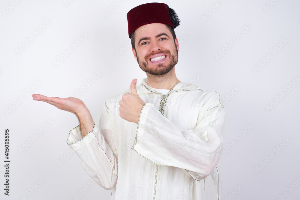 young handsome Caucasian man wearing Arab djellaba and Fez hat over white wall Showing palm hand and doing ok gesture with thumbs up, smiling happy and cheerful. young handsome Caucasian man wearing Arab djellaba and Fez hat over white wall Showing palm hand and doing ok gesture with thumbs up, smiling happy and cheerful.