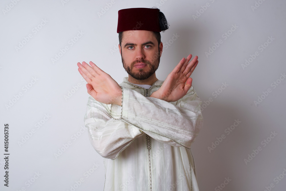 young handsome Caucasian man wearing Arab djellaba and Fez hat over white wall has rejection expression crossing arms and palms doing negative sign, angry face. young handsome Caucasian man wearing Arab djellaba and Fez hat over white wall has rejection expression crossing arms and palms doing negative sign, angry face.