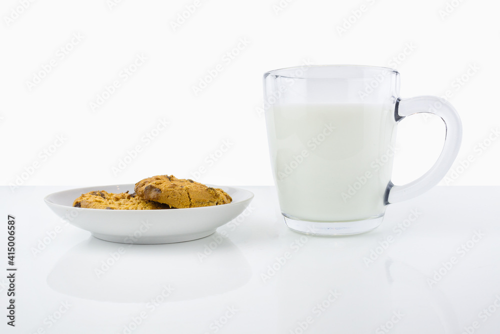 Two American cookies on white porcelain saucer and transparent cup of milk on white surface. Two American cookies on white porcelain saucer and transparent cup of milk on white surface.