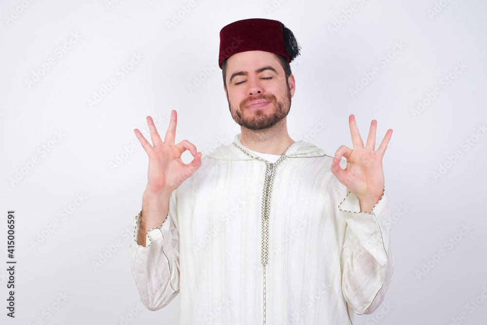 Naklejka premium young handsome Caucasian man wearing Arab djellaba and Fez hat over white wall relax and smiling with eyes closed doing meditation gesture with fingers. Yoga concept.