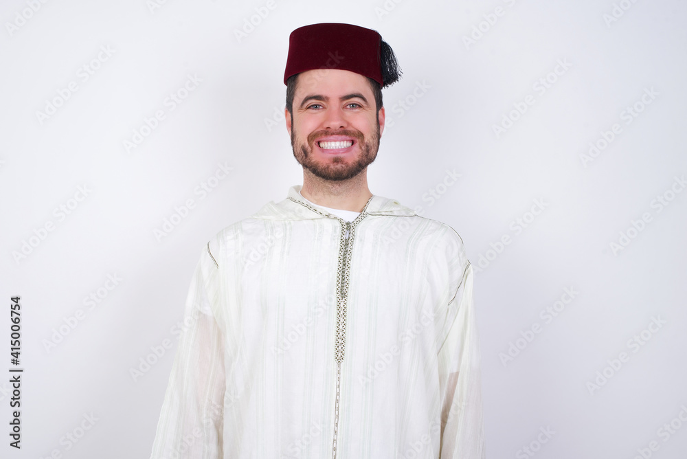 young handsome Caucasian man wearing Arab djellaba and Fez hat over white wall with a happy and cool smile on face. Lucky person. young handsome Caucasian man wearing Arab djellaba and Fez hat over white wall with a happy and cool smile on face. Lucky person.