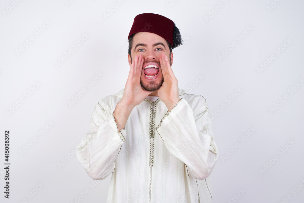 young handsome Caucasian man wearing Arab djellaba and Fez hat over white wall shouting excited to front. young handsome Caucasian man wearing Arab djellaba and Fez hat over white wall shouting excited to front.