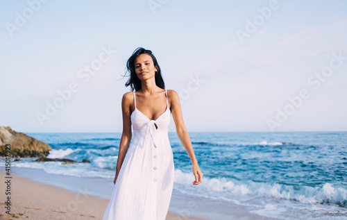Portrait of Latin woman dressed in comfortable white sundress walking at coastline beach during pastime near Caribbean sea  charming female model looking at camera during promenade on vacations