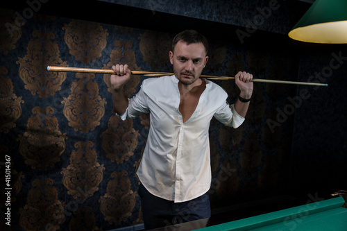 a man player stands at a billiard table holding a wooden cue with both hands and placing it on his shoulders behind his head