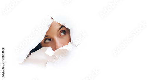 Portrait of a caucasian young woman looking through the hole in white paper. Distrustful look. Women's curiosity and gossip. A jealous wife. Spy concept.