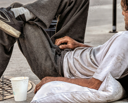 Homeless man with dirty tee shirt and sweatpants lying on sidewalk and pillow with paper cup panhandling 