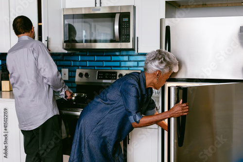 Wife getting drink from refrigerator while husband prepares to cook in kitchen, senior couple