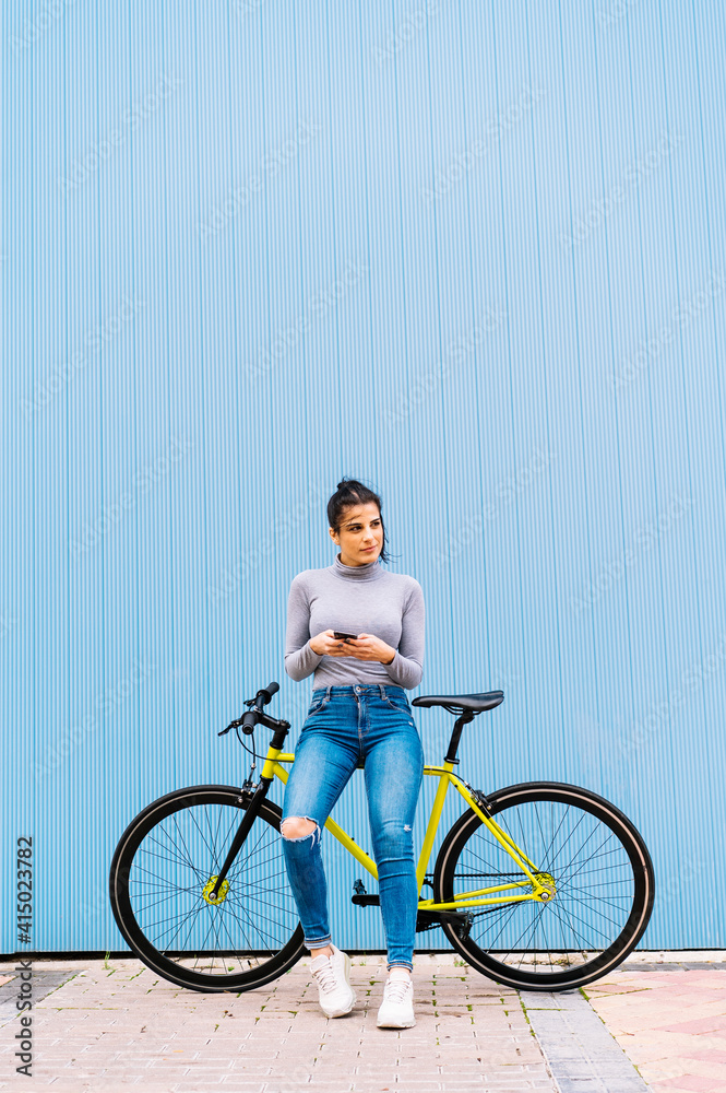 Woman using mobile phone looking away while sitting on fixie bike against blue wall Woman using mobile phone looking away while sitting on fixie bike against blue wall