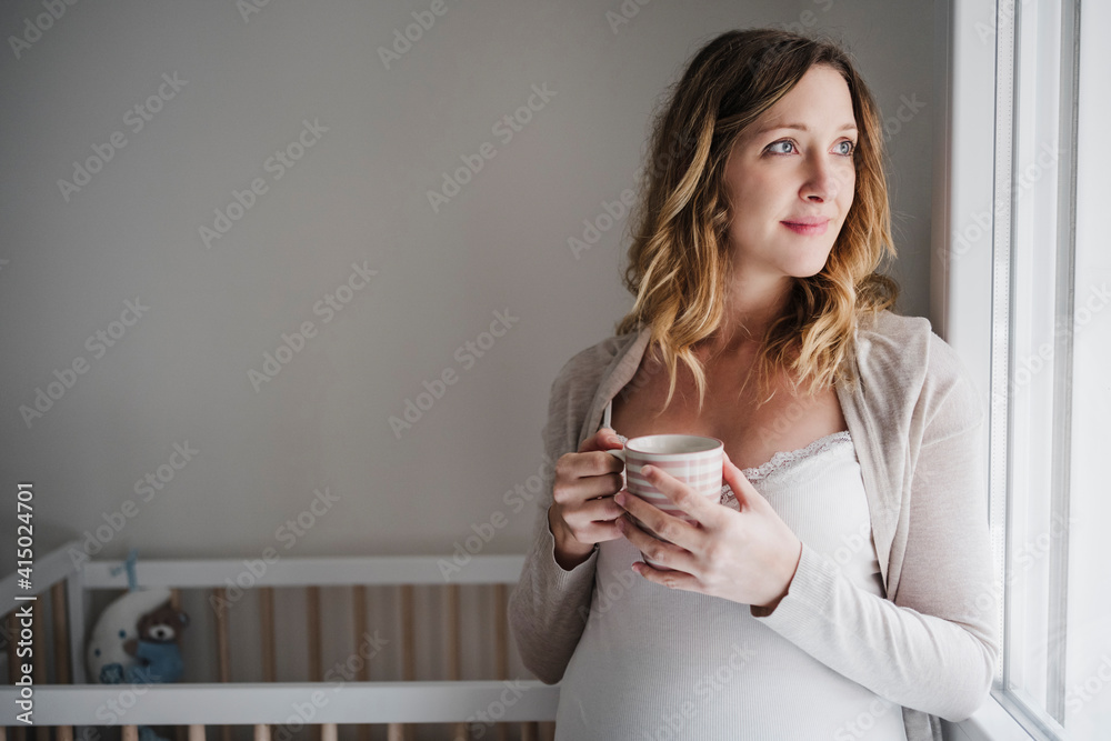 Thoughtful pregnant woman holding coffee mug while standing against crib at home Thoughtful pregnant woman holding coffee mug while standing against crib at home