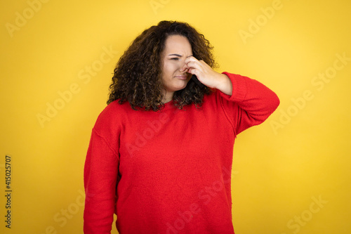Young african american woman wearing red sweater over yellow background smelling something stinky and disgusting, intolerable smell, holding breath with fingers on nose © Irene