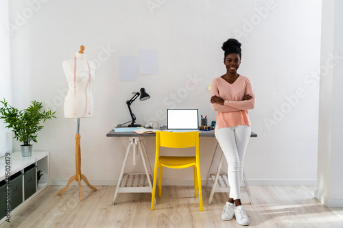Confident fashion stylist with arms crossed smiling while leaning on table at home photo