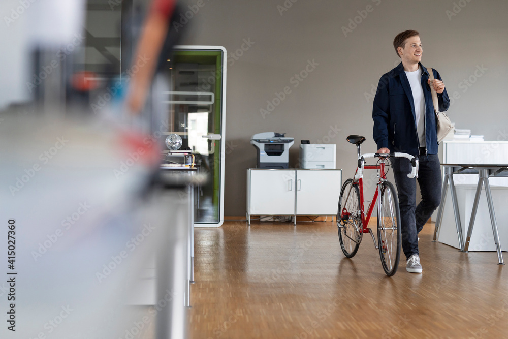 Businessman looking away while walking with bicycle at open plan office Businessman looking away while walking with bicycle at open plan office