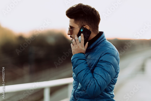 Young man in blue jacket talking on smart phone during sunset