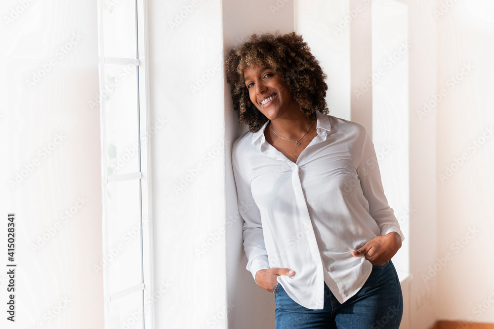 Mid adult woman with hands in pockets leaning on wall while standing by window at home Mid adult woman with hands in pockets leaning on wall while standing by window at home