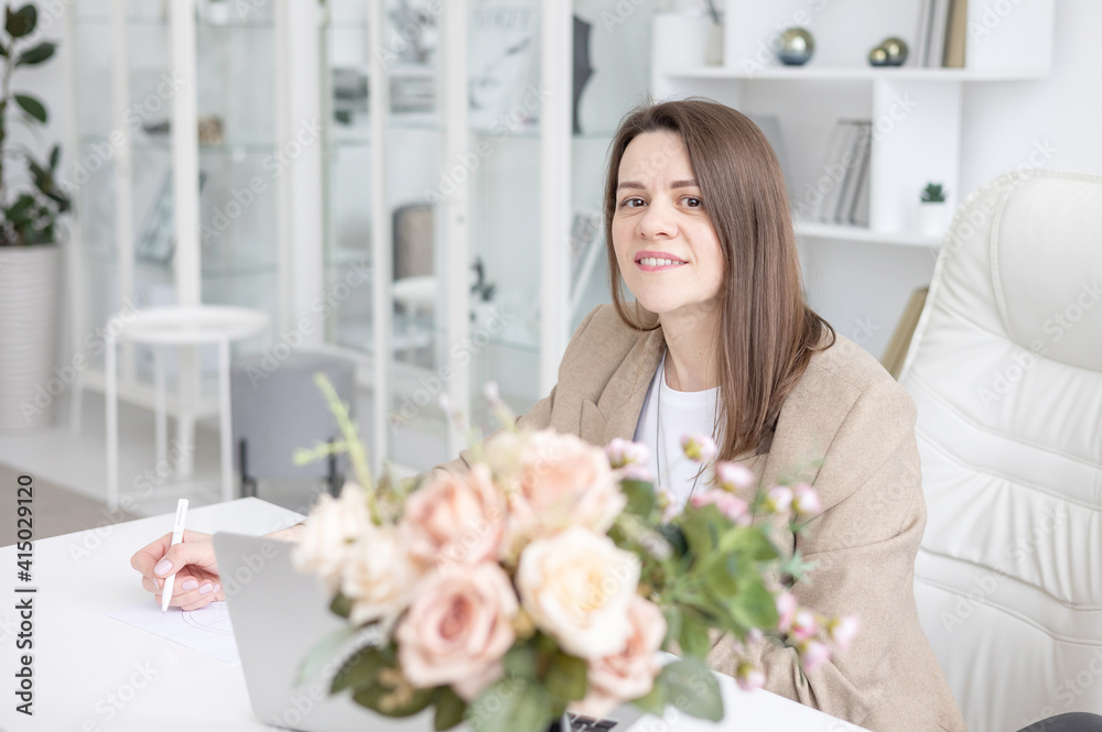 a young female coach works in a home office in a light interior with a laptop and flowers on the table, the concept of remote work