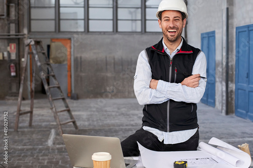 Male architect with arms crossed laughing while standing at table in building photo