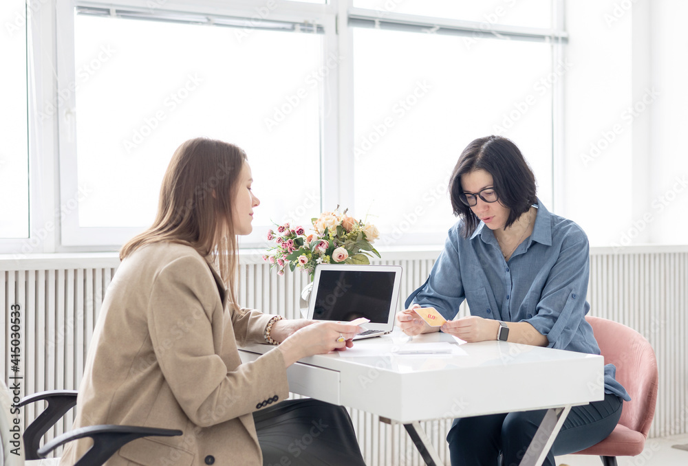 two young women conduct a coaching session in home office in light interior with laptop and flowers on tablework with metaphorical cards and psychological counseling two young women conduct a coaching session in home office in light interior with laptop and flowers on tablework with metaphorical cards and psychological counseling