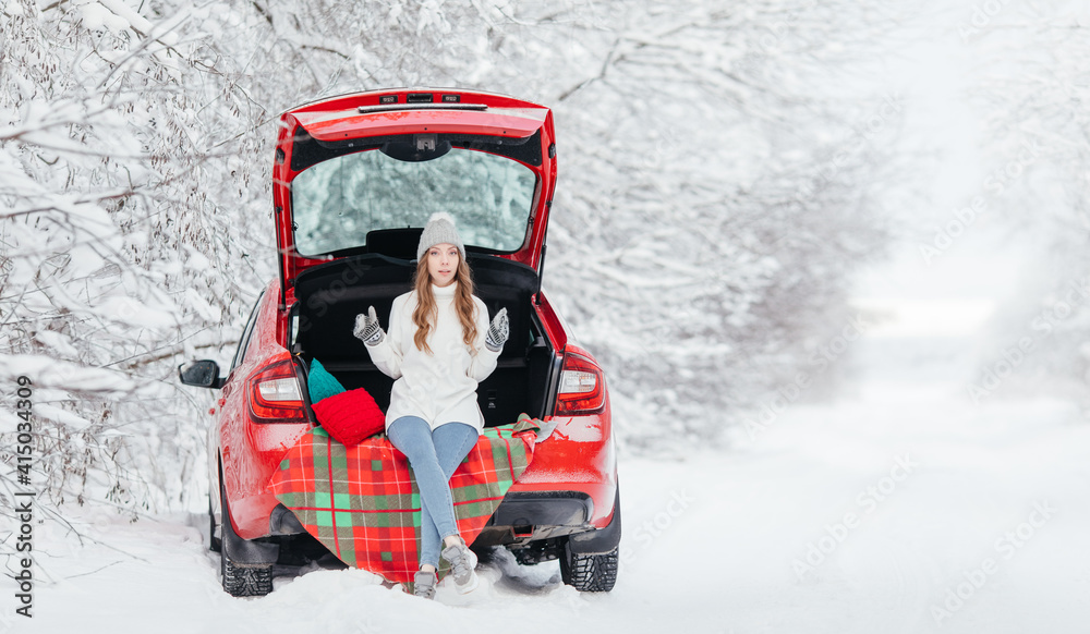 A woman with hot coffee in her hands sits in a red car on a snowy winter day in the forest.