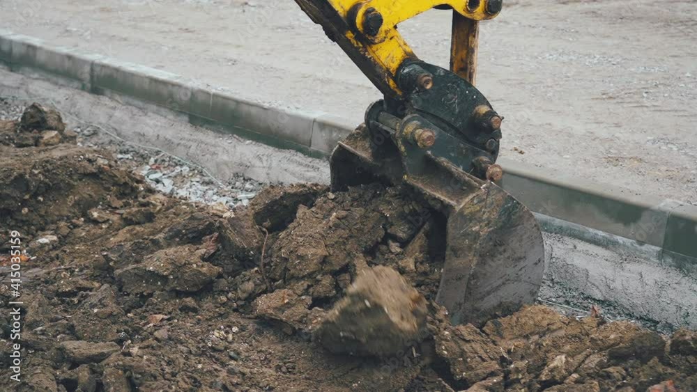 Excavator arm scoops up a bucket full of soil and rocks during roadworks in the countryside. Close up view of a big yellow excavator digging a deep trench near construction site Excavator arm scoops up a bucket full of soil and rocks during roadworks in the countryside. Close up view of a big yellow excavator digging a deep trench near construction site