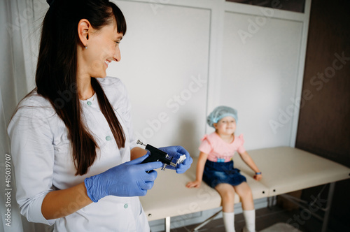 Young doctor holding a gun for piercing, smiling and looking towards the little girl