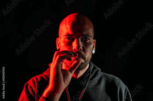 Serious male rocker smoking cigarette and looking at camera on black background in studio with red illumination