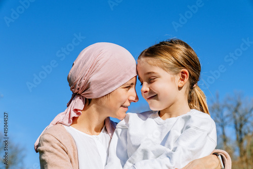 Happy mother with cancer wearing pink head scarf embracing little daughter on green park looking at each other