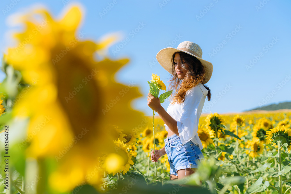 Side view of thoughtful ethnic female in hat holding flower standing in booming sunflower field and looking away Side view of thoughtful ethnic female in hat holding flower standing in booming sunflower field and looking away