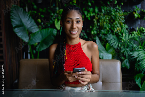 Portrait of attractive young afro latina with dreadlocks in a crochet red top posing with smartphone in restaurant table, Colombia