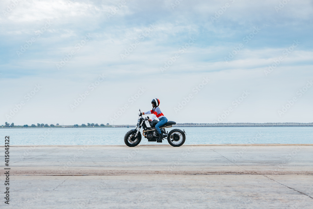 Side view of unrecognizable female biker in stylish clothes and helmet riding motorcycle on empty asphalt road near sea against cloudy blue sky Side view of unrecognizable female biker in stylish clothes and helmet riding motorcycle on empty asphalt road near sea against cloudy blue sky