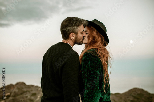 Side view of calm couple in stylish clothes and hat standing kissing at mountain seaside landscape on background of blue sea photo