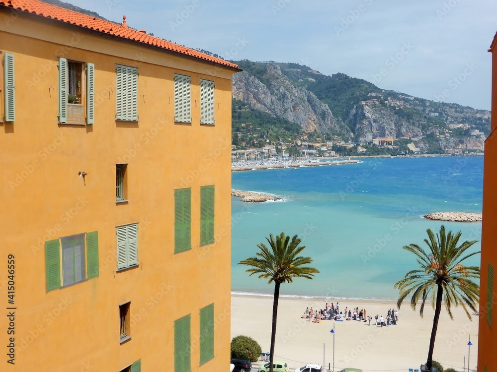Façade d'immeuble jaune dans la ville de Menton, avec vue sur la plage des Sablettes et l’eau bleu turquoise de la mer Méditerranée, sur la côte d'azur, dans les Alpes-Maritimes, en Provence (France)