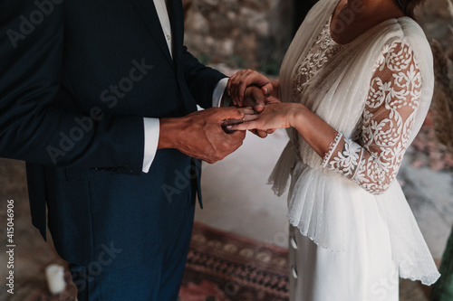 Side view of cropped unrecognizable ethnic groom putting ring on finger of bride in fancy wedding gowns holding hands gently with affection