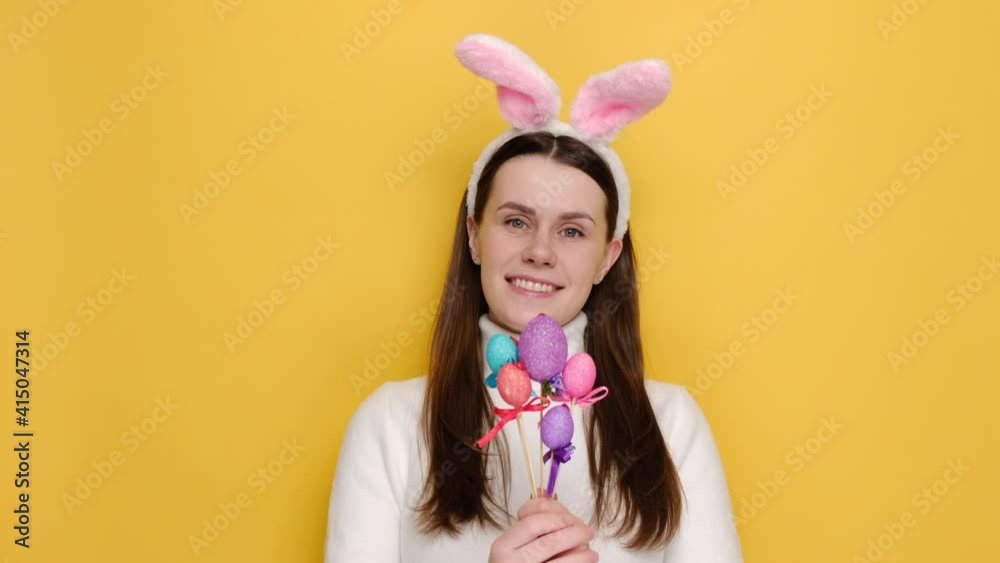 Portrait of smiling young girl 20s in pink bunny fluffy ears, suggests to take Easter egg, has symbolic thing in hands, wears white jumper, models over yellow studio background. Holidays concept Portrait of smiling young girl 20s in pink bunny fluffy ears, suggests to take Easter egg, has symbolic thing in hands, wears white jumper, models over yellow studio background. Holidays concept