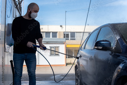 Lavado de coches. Un hombre joven limpiando su coche con agua a alta presión. Lleva una máscara KN95 ffp2 contra el SARS-CoV-2 photo