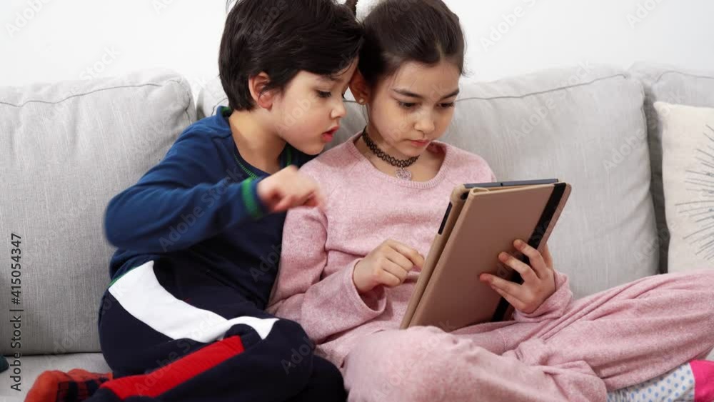 Two mixed-race kids sitting on sofa and looking with great interest into tablet. Real life, digital technology. 4K UHD