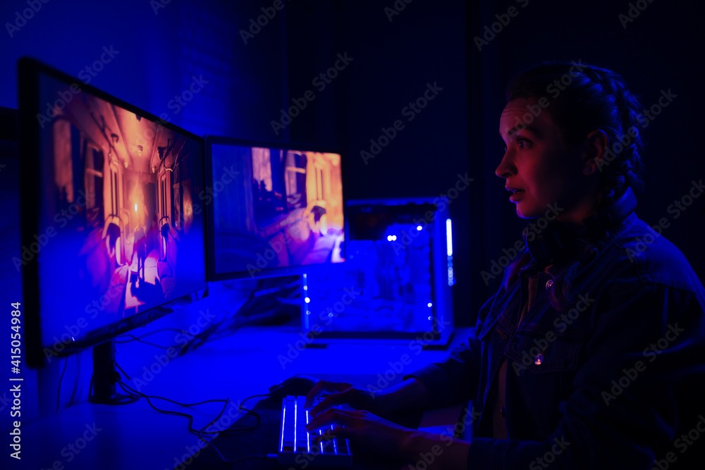 A girl with headphones plays a video game on a computer on large monitors. Gamer with mouse and keyboard. Online games with friends, win.e-sports, streams.Teenagers play adventure games.Neon lighting A girl with headphones plays a video game on a computer on large monitors. Gamer with mouse and keyboard. Online games with friends, win.e-sports, streams.Teenagers play adventure games.Neon lighting