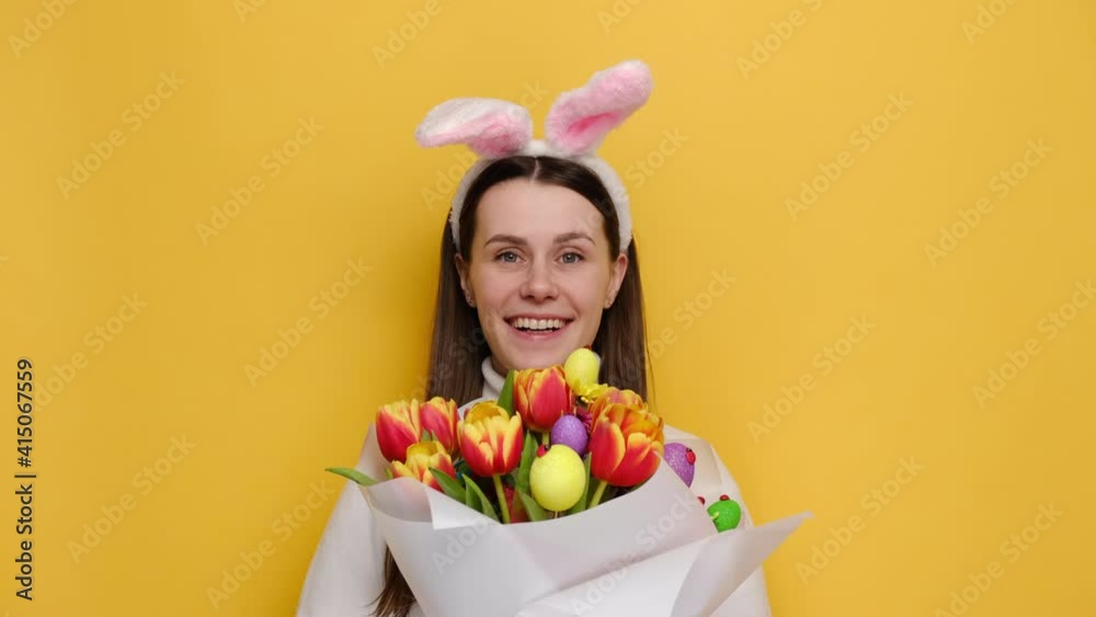 Sincere positive woman holding bouquet of fresh spring flowers decorated colorful small eggs, wears white jumper and bunny fluffy ears, smiles broadly feels happiness, isolated on yellow background Sincere positive woman holding bouquet of fresh spring flowers decorated colorful small eggs, wears white jumper and bunny fluffy ears, smiles broadly feels happiness, isolated on yellow background