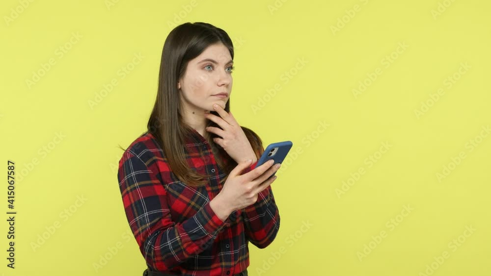 Serious concentrated business woman in checkered shirt typing on smartphone with thoughtful expression, working online, making notes. Indoor studio shot isolated on yellow background Serious concentrated business woman in checkered shirt typing on smartphone with thoughtful expression, working online, making notes. Indoor studio shot isolated on yellow background