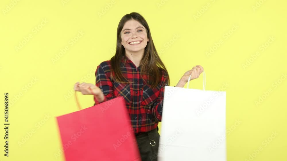 Smiling positive brunette woman in checkered shirt having fun and dancing holding in hands paper shopping bags, satisfied with successful purchases. Indoor studio shot isolated on yellow background Smiling positive brunette woman in checkered shirt having fun and dancing holding in hands paper shopping bags, satisfied with successful purchases. Indoor studio shot isolated on yellow background