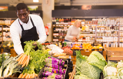 African male grocery worker in apron arranging fresh vegetables on shop counter