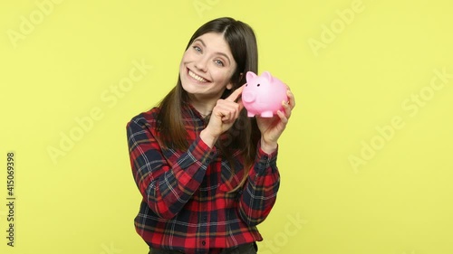 Smiling positive woman in checkered shirt holding piggy bank, suggesting you to put coins inside, saving money, deposit concept, banking. Indoor studio shot isolated on yellow background photo