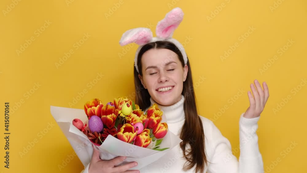 Cheerful woman in bunny fluffy ears hold bouquet of spring flowers decorated with Easter colored eggs enjoying good smell or pleasant fragrance, feel calm breathe fresh air, isolated on yellow wall Cheerful woman in bunny fluffy ears hold bouquet of spring flowers decorated with Easter colored eggs enjoying good smell or pleasant fragrance, feel calm breathe fresh air, isolated on yellow wall