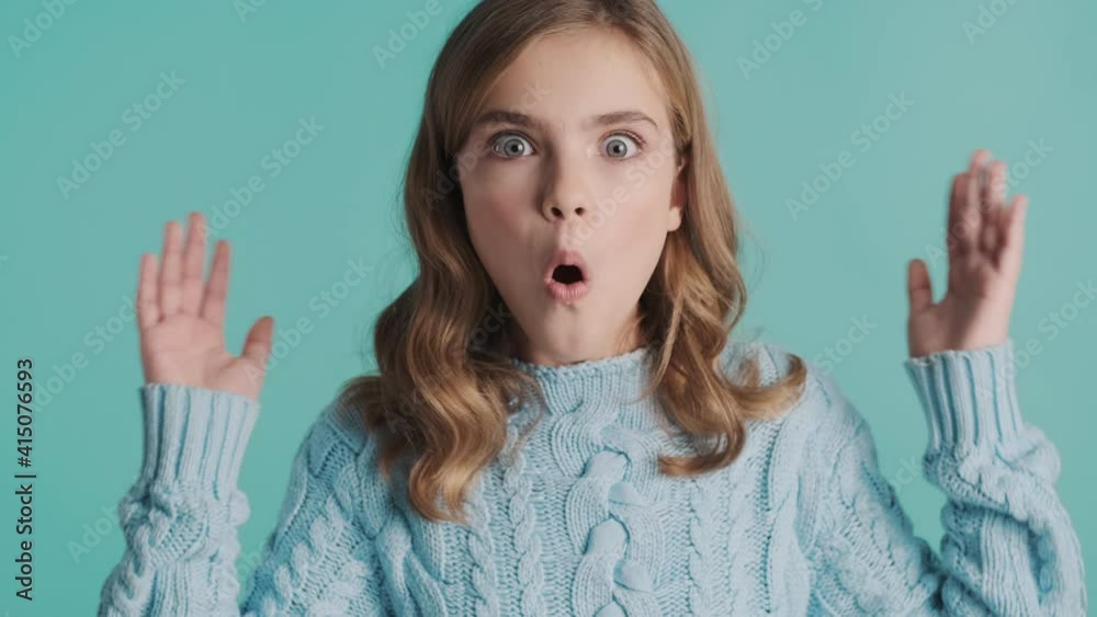 Amazed blond teenage girl in cozy sweater looking shocked on camera isolated on blue background. Wow expression