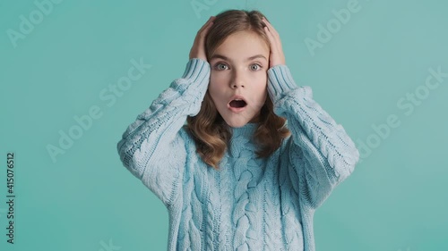 Shocked blond teenager girl in cozy knitted sweater looking astonished in camera over blue background. Wow face expression