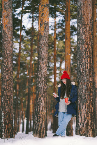 Caucasian girl in red hat with paper cup in the forest looking at her phone. Walk in the snowy forest with coffe to go