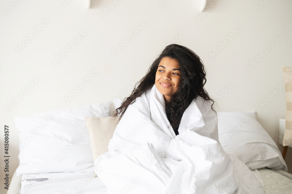 Young African American teenage girl with Afro hairstyle covered in a cozy white soft blanket staying at home in the warm bed, looking in the window, feeling happy and blessed, good morning concept Young African American teenage girl with Afro hairstyle covered in a cozy white soft blanket staying at home in the warm bed, looking in the window, feeling happy and blessed, good morning concept